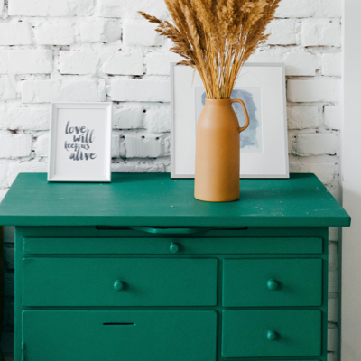 a photo of a blueish green dresser sitting against a white brick wall with picture frames and a vase holding a plant sitting on top of it